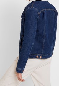 Person med mörkblå denimjacka med vit sherpa-krage och krämfärgade byxor, stående mot en enkel ljusgrå bakgrund.