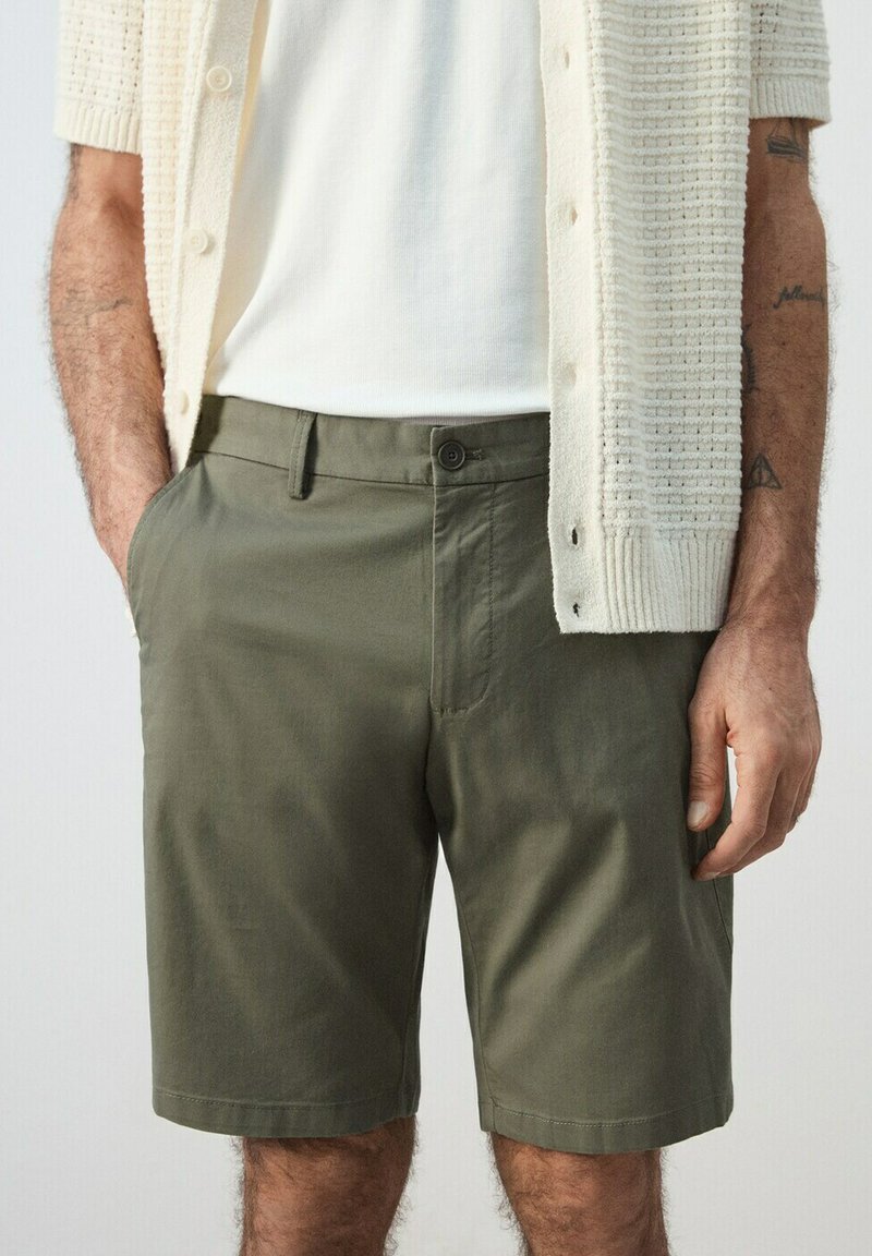 Homme portant un short vert olive sur mesure avec poches latérales, une chemise blanche et un cardigan crème texturé avec des boutons, debout les mains détendues.