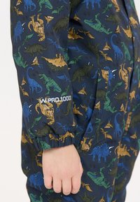 Dino-patroon waterdichte overall in donkerblauw met groene, bruine en blauwe illustraties; voorzien van elastische manchetten en pocketdetails.