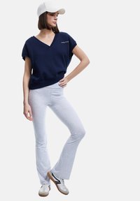 Kvinna iklädd en marinblå V-ringad topp, ljusgrå utsvängda leggings, vit keps och vita sneakers med gummisula, poserar med handen på höften.