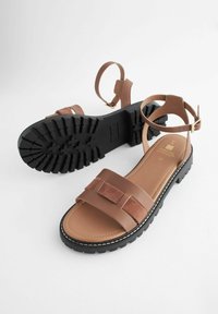 Brune sandaler med et struktureret ruskindsrem og glat læderbetræk. Tyk sort gummi sål med et robust mønster og spænde-lukning.