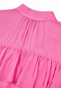 Kevyt, kudottu vaaleanpunainen paita, jossa on korkea kaulus ja laskostettu takakappale. Helmassa on rypytetty muotoilu lisäämään tekstuuria.