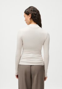 Top de manga larga en tejido beige claro con un diseño ajustado, que presenta un cuello alto y una textura suave, combinado con pantalones grises holgados.