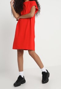 Robe rouge à manches courtes avec une coupe décontractée, fabriquée en tissu lisse. Portée avec des baskets noires et des chaussettes blanches pour un look décontracté.