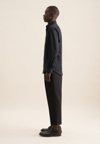 Camicia nera con maniche lunghe, colletto piatto e texture liscia, abbinata a pantaloni e scarpe neri, vista di lato.