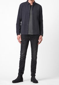 Tmavá košile s knoflíky, šedé tričko pod ní, roztrhané černé džíny a černé kotníkové boty. Tkanina vypadá měkce a má ležérní střih.