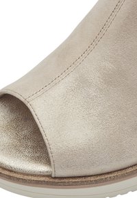 Zapato abierto de punta en color beis, texturizado y con un suave brillo metálico. Cuenta con una costura cosida y una suela sólida de color claro en contraste.