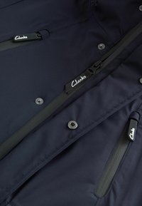 Chaqueta azul marino hecha de tela duradera, con múltiples bolsillos con cremallera y botones metálicos a presión, con la marca Clarks.