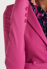 Osoba nosící jasně růžové sako se čtyřmi stejnobarevnými knoflíky a květinovou halenku s fialovými, modrými a růžovými tóny, částečně viditelnou.