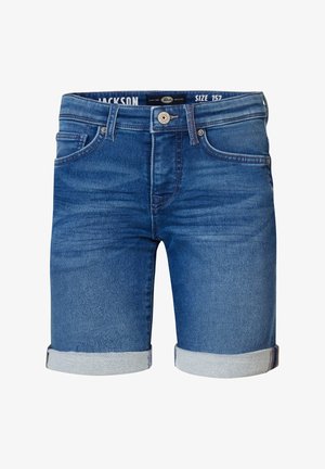 Stehende Person mit pfirsichfarbenem Kurzarm-T-Shirt, blauen aufgerollten Jeansshorts, weißen Socken und weißen Sneakern vor einfachem Hintergrund.