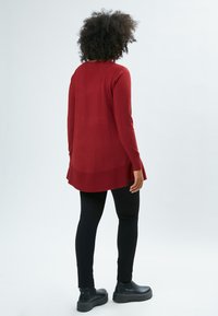 Langärmliger, roter Strickpullover mit einem abgerundeten hinteren Saum, getragen über schwarzen eng anliegenden Leggings und kombiniert mit schwarzen Plateaustiefeletten.