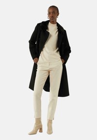 Black wool coat with a fur collar, beige knit sweater, cream tailored pants, and beige high-heeled boots, standing against a neutral background.