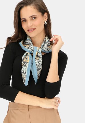 Kobieta w czarnym topie, beżowych spodniach, srebrnych kolczykach i niebieskim szaliku w wzory, wiązanym wokół szyi, uśmiechająca się z dłonią blisko twarzy.