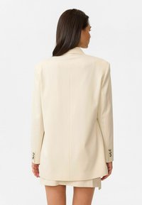 Beige oversized blazer met schoudervulling, enkele achterventilatie en knoopmanchetten. Gladde stof met een getailleerde pasvorm en een minimalistisch ontwerp.