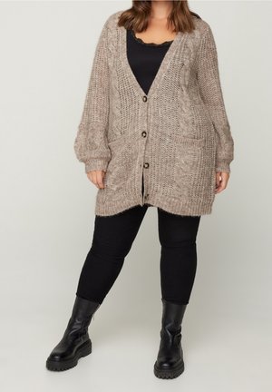 Nainen pukeutunut ylisuurikokoiseen beigeen palmikkoneuleiseen villatakkiin, mustaan toppiin, mustiin leggingsseihin ja mustiin paksupohjaisiin nahkasaappaisiin, seisoo jalat leveässä haara-asennossa.