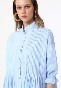 Chemise boutonnée bleu clair avec des rayures verticales, design plissé, coupe oversized et manches froncées. Présente un col montant et des boutons bleus.