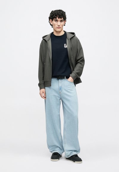 Σκούφο σκούρο πράσινο πάνω από μπλε πουλόβερ, ανοιχτό μπλε τζιν με φαρδύ πόδι και μαύρα αθλητικά παπούτσια. Απλές υφές και ελάχιστες λεπτομέρειες σε όλο το σύνολο.