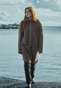 Brun fleecejakke med høj krave og lynlåsfortil. Lange ærmer, sidelommer og en tekstureret overflade. Sorte bukser og støvler fuldender outfittet.