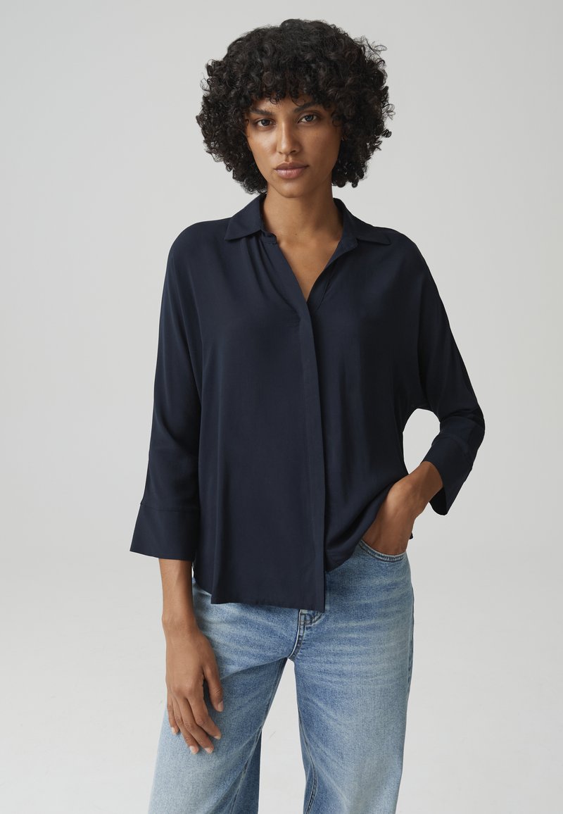 Blusa blu navy a maniche lunghe con collo a camicia e vestibilità ampia. Tessuto morbido con una texture liscia, abbinata a jeans azzurri.
