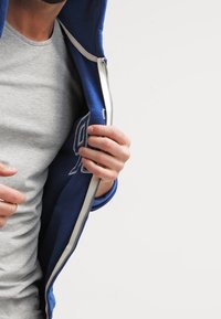 Uomo che indossa una t-shirt grigia apre una felpa blu con zip non chiusa, dotata di un interno bianco e una toppa con il numero nove sul lato sinistro.