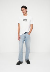 Biała bawełniana koszulka z czarnym logiem "Calvin Klein", w połączeniu z jasnoniebieskimi dżinsami i czarnymi trampkami. Prosty, luźny krój.