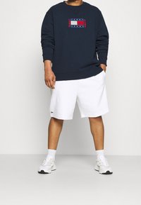 Granatowa bluza z kapturem z logo na piersi, zestawiona z białymi szortami. Model ma na nogach białe trampki i gładkie, niewidoczne skarpetki.