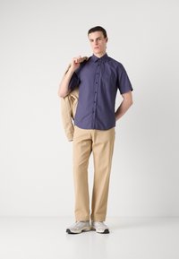 Korte ermers marineblå skjorte med brystlomme, beige bukser og grå sneakers. Modellen holder en beige jakke over den ene skulder, poserer oppreist.
