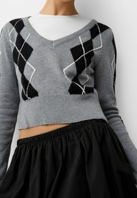 Grå croppad tröja med V-ringning, med ett svart och vitt argyle-mönster. Tyget ser mjukt ut och har ribbade muddar och nederkant. Matchas med en svart kjol.