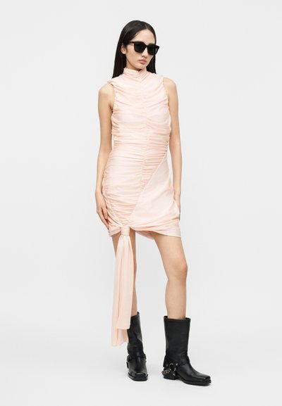 Γυναίκα με ρούχα ρουστίκ, αμάνικο παλ παστέλ ροζ φόρεμα με δεμένη γάζα, μαύρες αστραγαλομπότες με αλυσίδες και μαύρα γυαλιά ηλίου, στέκεται μπροστά σε λευκό φόντο.