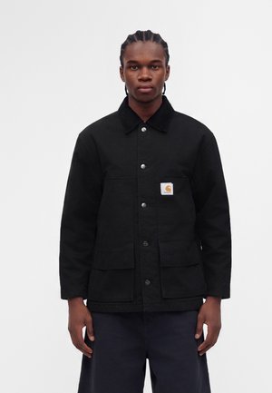 Jonge man met gevlochten haar die een zwarte, geknoopte jas draagt met grote zakken en een klein vierkant logo op de borstzak.