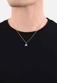 Collana in oro con un piccolo pendente "M" texture, indossata sopra una maglietta nera a girocollo su uno sfondo neutro.