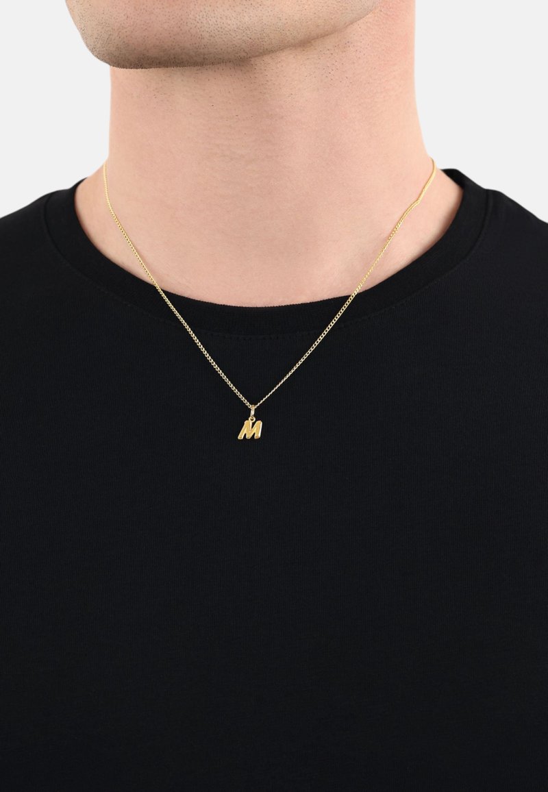 Collana in oro con un piccolo pendente "M" texture, indossata sopra una maglietta nera a girocollo su uno sfondo neutro.