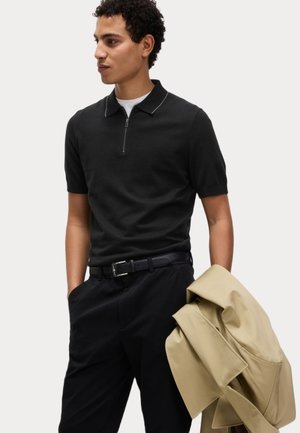 Polo nero a maniche corte con zip, abbinato a pantaloni neri e una giacca beige chiaro appoggiata sul braccio, presenta una texture liscia.