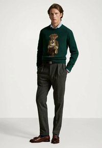 Νεαρός άνδρας με σκούρο πράσινο πουλόβερ με γραφικό αρκουδάκι, γκρίζες πλισέ παντελόνες, καφέ δερμάτινες μοκέτες, στέκεται με τα χέρια στις τσέπες.