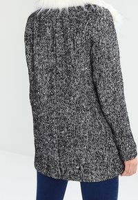 Abrigo texturizado en blanco y negro con cuello de piel, mangas largas y un diseño a la altura de la rodilla, que presenta un sutil patrón de espiga.