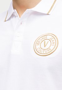 Λευκό πόλο πουκάμισο με γιακά διακοσμημένο με χρυσή λεπτομέρεια και χρυστό κεντημένο κυκλικό λογότυπο Versace Jeans Couture στο στήθος.