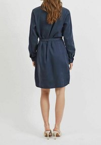 Mörkblå skjortklänning med långa ärmar, bälte i midjan och knälång fåll. Slät textur och minimalistisk design.