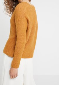 Kvinna som bär en senapsgul ribbad stickad tröja med långa ärmar och vita byxor, visad från sidan mot en vit bakgrund.