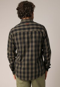 Man met krullend haar, gekleed in een groen en zwart geruite langste mouw overhemd, staat met zijn rug naar de camera tegen een effen achtergrond.