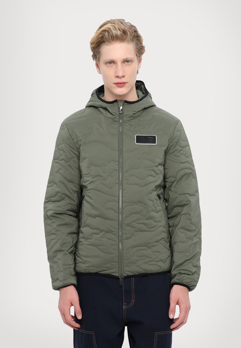 Quilted olive green jacket with a hood, zip closure, and a black logo patch. Features side pockets and elastic cuffs. Textured surface.