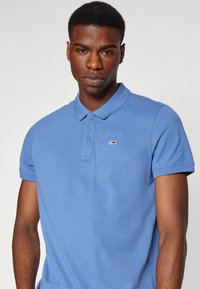Camiseta polo azul de algodón, con un cuello clásico, tapeta de dos botones y un pequeño logo en el lado izquierdo del pecho. Mangas cortas.