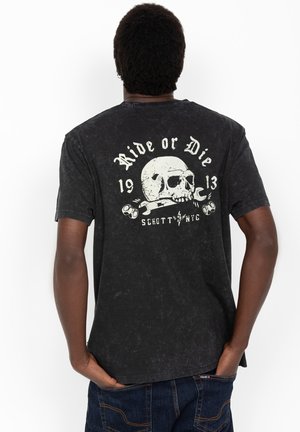 Mann trägt schwarzes T-Shirt mit Totenkopf, Schraubenschlüssel und dem Text „Ride or Die 1913 Schott NYC“ auf dem Rücken, Hände in den Taschen, vom Betrachter weggewandt.