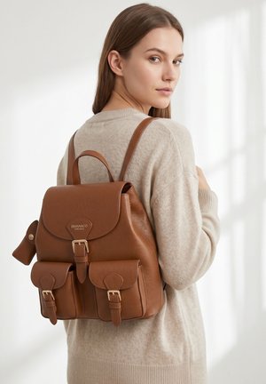 Jonge vrouw met beige trui draagt een bruine leren rugzak met gespen en twee voorvakken, zijwaarts tegen een witte achtergrond.