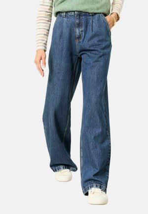 Person, die lockere blaue Jeans, weiße Sneakers, ein grünes Top und ein gestreiftes Langarmshirt trägt, mit einer Hand in der Tasche.