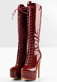 Bottes en cuir verni rouge brillant jusqu'aux genoux, avec une plateforme haute et un talon stiletto. Dotées d'un laçage à l'avant avec des œillets métalliques.
