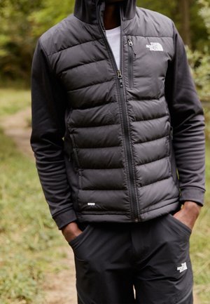 Person iført en sort puffer vest og sort langærmet skjorte står udendørs med hænderne i lommerne på en natursti.