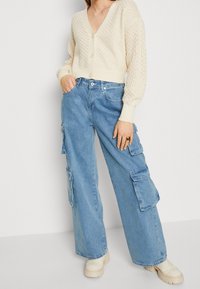 Ljus beige kabelstickad cardigan med djup V-ringning och knappar, tillsammans med ljusblå vida cargobyxor med sidofickor. Krämfärgade kraftiga skor.