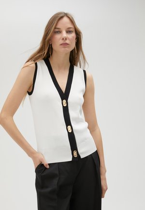 Weißer ärmelloser Cardigan mit tiefem V-Ausschnitt, schwarzem Besatz und einer mittigen Reihe goldfarbener Knöpfe, kombiniert mit schwarzen Hosen.