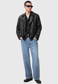 Schwarze Leder-Bikerjacke mit breiten Revers, Reißverschluss-Taschen und taillierter Passform; kombiniert mit hellblauen Straight-Leg-Jeans und schwarzen Schuhen.