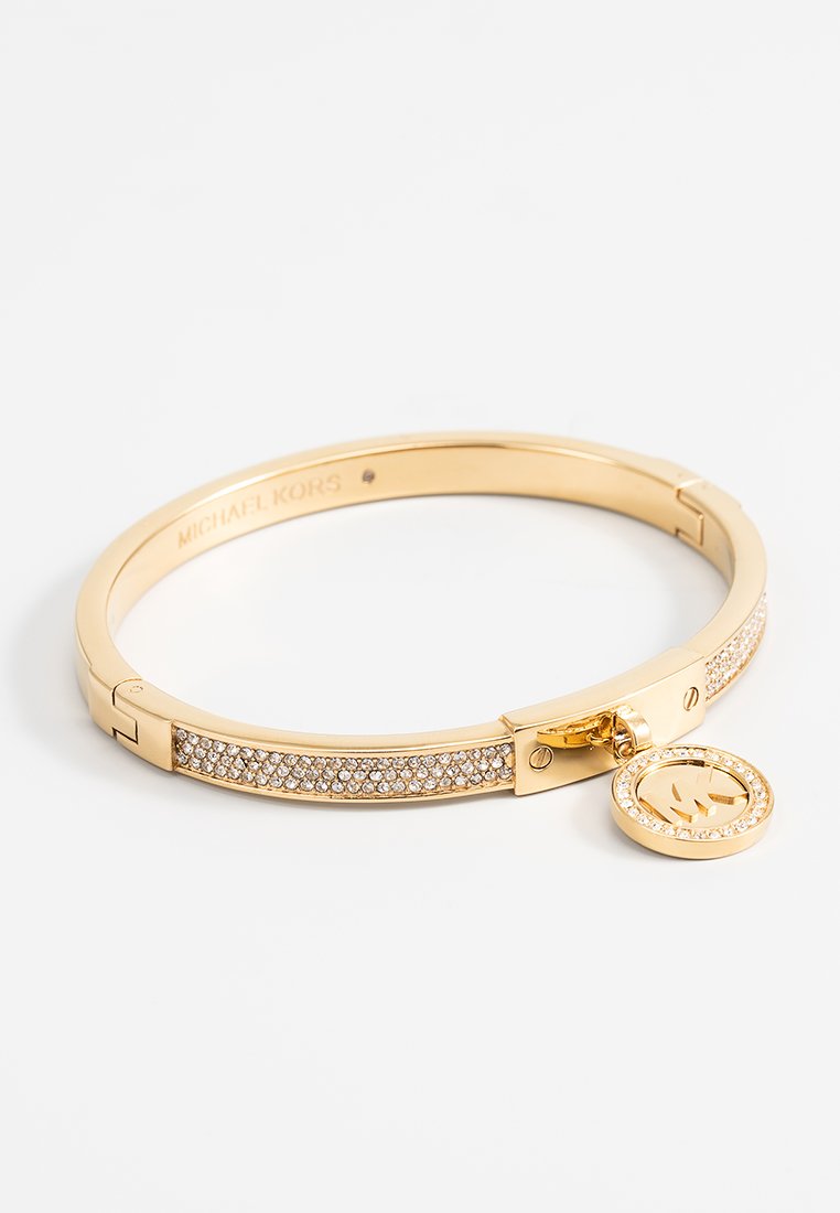 Bracelet jonc en tone doré avec une rangée de cristaux transparents, une fermoir à clip, et un charm logo suspendu. Fini lisse et poli.